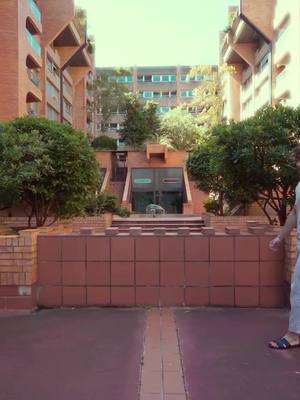 An outdoor shot of a courtyard with buildings, trees and a person walking in the scene. The pathway and foreground is a dark reddish color made of concrete. In the center is a block wall with an opening. There is a tall building to the left and right side of the image, the top of the buildings have trees. The sky is a light color, almost white, it appears a cloudy day.