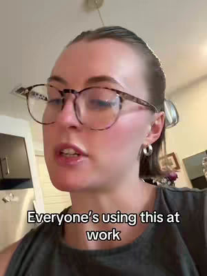 A woman with fair skin and light-colored hair is centered in the frame, wearing glasses with a dark tortoiseshell pattern, and a gray tank top. The background is a room with a light-colored ceiling and walls, and an overhead light fixture is visible. Text in a black box reads: "Everyone's using this at work".