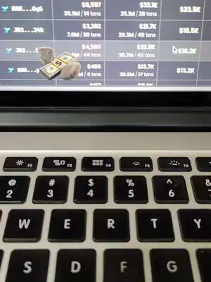 A close-up shot of a silver laptop keyboard with a screen displaying financial data in the background. The screen shows a table of numbers relating to financial transactions. An image of money is layered on the laptop screen. The foreground is filled with the black keys of the laptop keyboard, with white lettering.