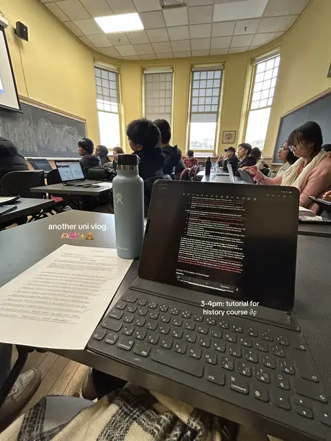 The image is a first-person point of view inside a university classroom. A laptop with a keyboard is in the foreground, with an open document and a keyboard on the desk. To the right of the laptop is a water bottle and a sheet paper with handwritten notes. In the background other students are visible seated at desks.