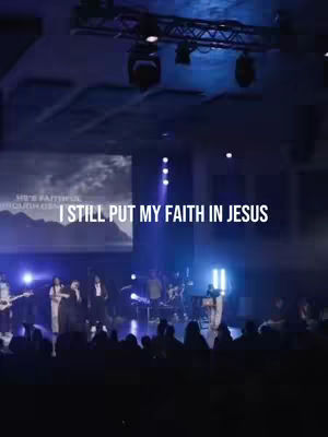 A stage is lit with blue and white lights, with a band and choir performing in front of a large screen. Text overlayed says, "I still put my faith in Jesus."