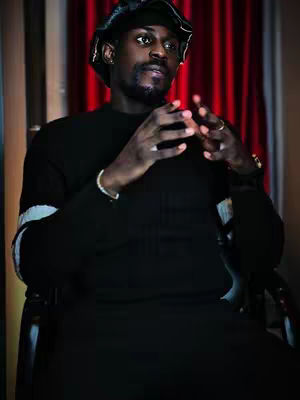 A well-lit medium shot features a well-dressed man seated, hands clasped together. He wears a black top and cap, with a red curtain in the background.