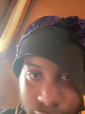 A close-up selfie featuring the upper body of a person wearing a dark, possibly black, head covering. The individual's face is the primary focus, with the eyes prominently visible. Soft lighting suggests an interior setting, and the color palette is dominated by dark tones.
