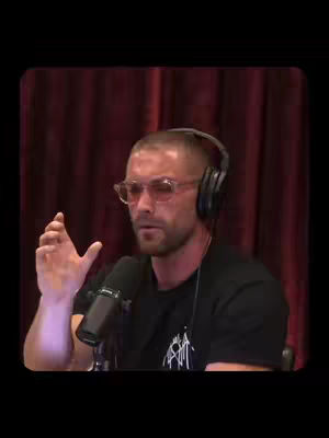 A man with a shaved head, wearing glasses, a black shirt, and headphones gestures with his right hand while speaking into a microphone. He is seated in front of a red curtain.