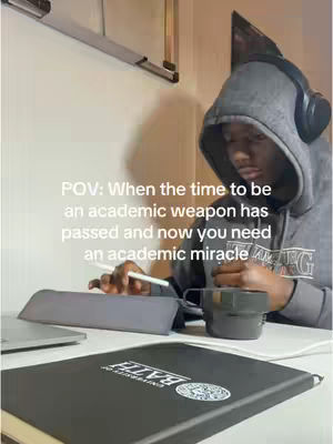 A person wearing a grey hoodie and headphones is sitting at a desk, looking down at a notebook and tablet. There's a coffee mug to the right. Text overlay says 'POV: When the time to be an academic weapon has passed and now you need an academic miracle.'