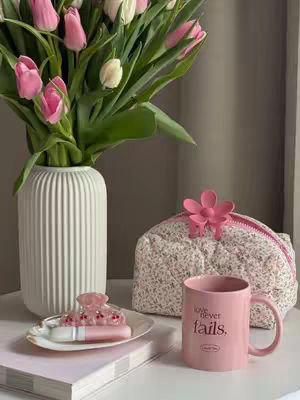 An indoor, still-life shot featuring a bouquet of pink and white tulips in a white fluted vase, a pink coffee mug, a floral patterned pouch, and a plate holding a hair clip and lip gloss. The color palette is soft and pastel, with the main colors being pinks, whites, and light greens. The arrangement is placed on a white surface, likely a table or shelf, against a neutral light gray background. The mug has the text 'love never fails' on it.