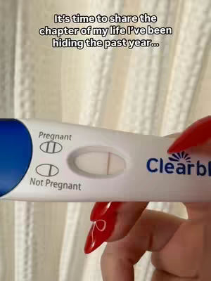 A close-up shot of a hand holding a Clearblue pregnancy test. The test shows a faint second line, indicating a possible pregnancy. The background is a neutral, blurred setting, likely a bathroom or bedroom.
