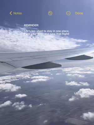 The image shows an airplane wing, with the wing extending across the top third of the frame. The sky is a vibrant blue, with fluffy white clouds scattered throughout. The angle suggests a view from inside the plane, looking out at the wing and the sky. A notepad is superimposed containing a reminder.