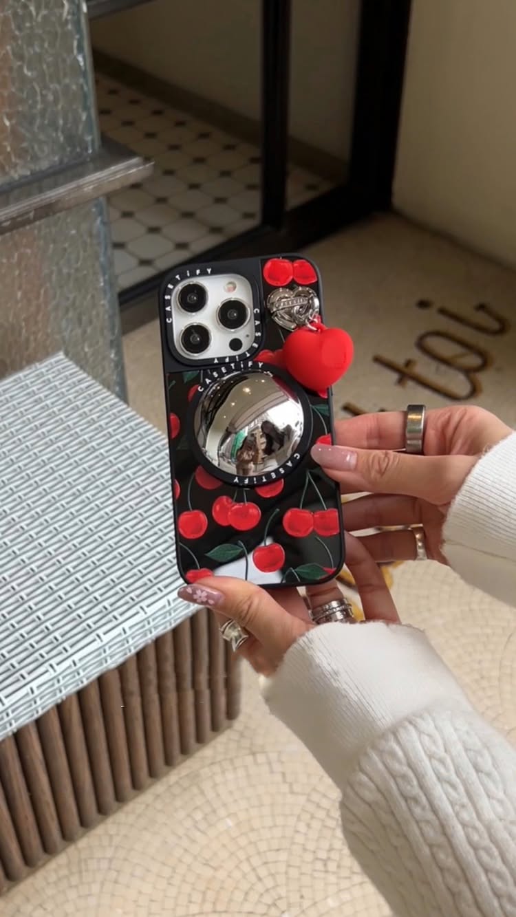 A person's hands holding a black phone case with a cherry pattern and a fish-eye mirror grip stand. The person is wearing a white sweater and rings on their fingers. The background includes a mosaic tile floor and a window.