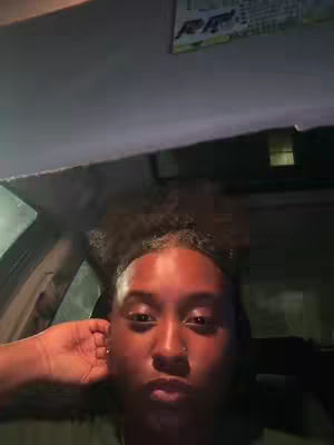 A selfie-style shot taken inside a vehicle, with the main focus on the person's face. The lighting is somewhat dim, suggesting it's either nighttime or a shaded environment. The person has their right hand up near their head. The color palette mainly consists of dark tones with the main subject's skin being the brightest point of focus.