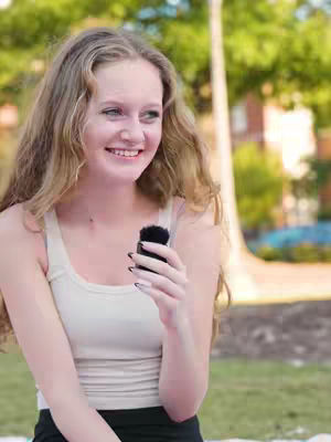 A young woman with long, wavy blonde hair is smiling and looking slightly off-center to her left while holding a small black microphone in her right hand. She is wearing a cream-colored tank top and a black bottom. The background includes elements of a park with greenery and trees.