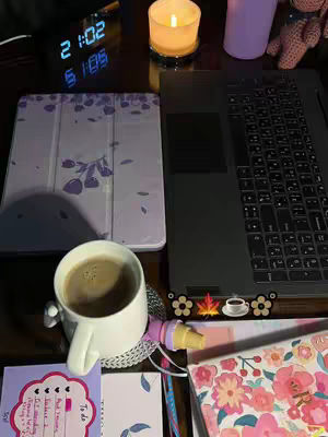 A dimly lit desk with a laptop, tablet, coffee cup, candle, and study materials. The overall composition is warm and inviting, creating a study atmosphere.