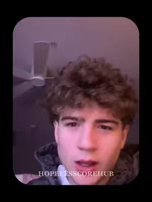 A close-up shot of a young man with curly brown hair looking towards the camera with a slightly open mouth and a neutral expression. The background includes a ceiling fan and a light purple-toned wall.
