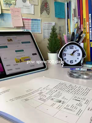 A desk setup with school supplies, a laptop, and a clock. The laptop screen displays a website with colorful graphics. The clock is black and white, with the hands pointing to about 10:10. There's a small, artificial Christmas tree, books, and papers. The image is well-lit, with a focus on the workspace, symbolizing productivity.