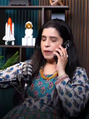 A woman with long dark hair sits in a room, holding a phone to her ear. She wears a patterned dress and a necklace. A microphone is in front of her. The background includes a wooden shelf with decorations.