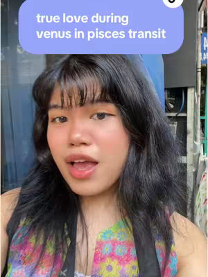 A selfie-style shot of a woman with dark hair and a colorful top. Above her head, a text overlay reads, "true love during venus in pisces transit" in a rounded rectangle. The background is blurred, but appears to be outdoors with some buildings and possibly foliage visible.