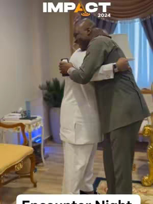 Two men embrace, one in a suit, the other in white. The scene is indoors, with a blurred background and text overlays. The image has a vertical orientation.