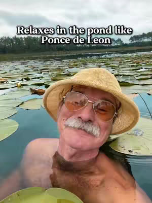 An older man with a mustache and sunglasses is in a pond filled with lily pads. The text overlay reads 'Relaxes in the pond like Ponce de Leon'.