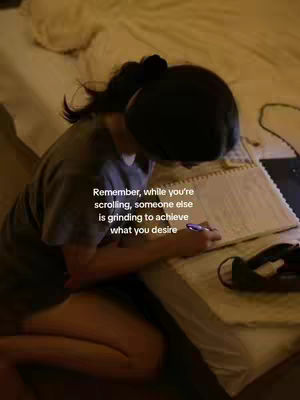 A person is sitting on a bed, angled away from the camera, studying. The overall tone is warm, with soft brown and yellow lighting coming from a soft, warm source. The person is wearing a gray t-shirt, has dark hair pulled back, focused on writing on a notepad that sits in front of them. The composition shows a bed in the background.