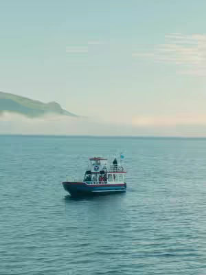 An eye-level shot of a boat in the midle of the sea with a misty background, The boat is white and red with a dark blue bottom, there's a mountain, also partially obscured by mist, at the back.