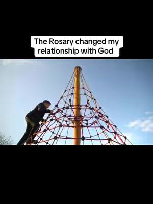 The cover image shows a person climbing a climbing frame made of a yellow pole and a web of red and black ropes. Text in white rectangular box says 'The Rosary changed my relationship with God'. The background is a blue sky with some clouds.