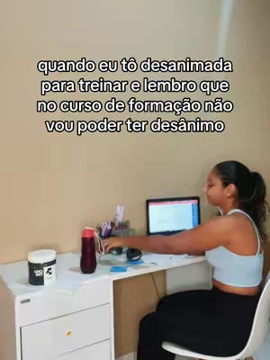 A woman is sitting at a desk with a laptop, a water bottle and a protein container are visible. On the laptop the screen is on. The woman is working. The background is a wall and also a table