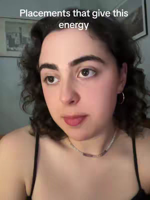 A young woman is centered in a close-up shot, looking slightly off-camera with a subtle expression. The framing highlights her face, with dark, curly hair framing her face. She is wearing a black top and a necklace. The background is a room with a painting and dim lighting.