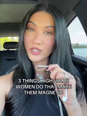 A woman with long, dark hair is positioned in the driver's seat of a car. She is holding white earbuds in her right hand. Text overlayed on the image reads, '3 THINGS HIGH VALUE WOMEN DO THAT MAKE THEM MAGNETIC'. The background shows a view of greenery and water.