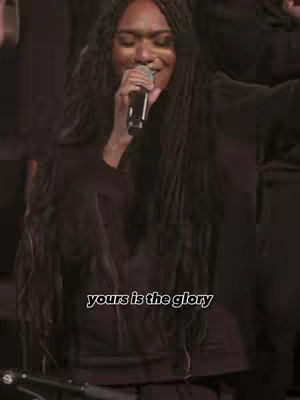 A woman with long, dark braids sings into a microphone. The background is dark, possibly a stage setting. Text overlayed at the bottom reads 'yours is the glory'.