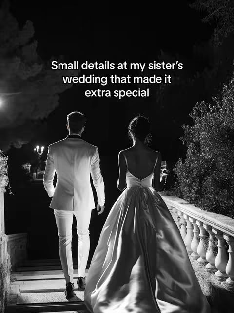 A black and white photo depicts a well-dressed couple walking away from the camera on a set of steps. The man, dressed in a white suit, leads the way, while the woman, in a flowing wedding gown, follows behind him. The background fades into darkness, with a few trees and architectural elements visible. The overall composition is elegant and suggests a special event.