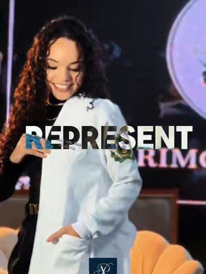 A woman with curly brown hair smiles broadly while partially covered by a white lab coat. The word "REPRESENT" is overlaid on the coat in bold white letters. The background suggests an auditorium or stage, with subdued lighting and a partially visible circular graphic element. An orange chair is visible at the bottom.