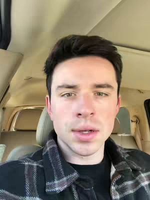 A man is seated in a car, facing the camera. He has short, dark hair and is wearing a black and plaid button-down shirt. The car's interior is visible, including the headrests. The lighting is natural, and the man seems to be in a casual setting.
