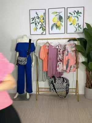 The image shows a woman's outfit represented on a mannequin with colorful dresses and tops on a clothing rack, and three framed artwork of lemons. A woman in a pink shirt is partially visible in the foreground. The room has a light-colored floor and white walls.