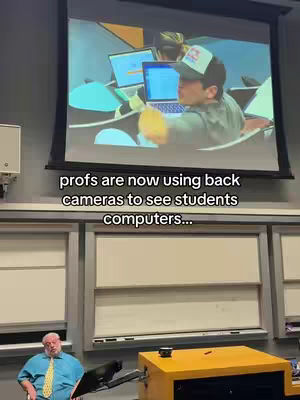The image shows a classroom setting. A large screen displays a projected image of a person using a laptop. Below, multiple rows of windows and blinds provide the background, while a teacher is sitting on the left, looking to the side.