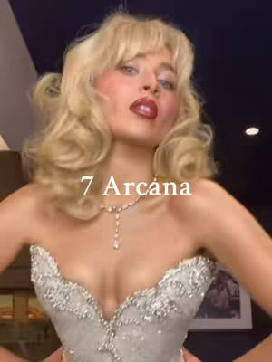 A woman with blonde, wavy hair is shown from the chest up. She wears a white, embellished dress, a diamond necklace, and has red lipstick on. The text "7 Arcána" is overlaid on the image.