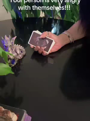 The image features a hand holding a deck of cards, with text overlayed at the top. The background is dark and reflective, with a glimpse of green foliage. A small decorative dragon is visible on the left, and more cards are at the bottom. The lighting appears to be from above.