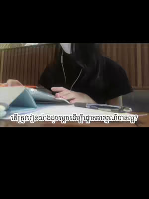 A person is seated at a table, seemingly studying with a notebook and pen in front of them. They are wearing a face mask and earphones. The background has wooden paneling.