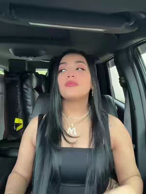 A woman with long black hair is seated inside a car, looking upwards with a slight pursing of her lips. A necklace with a letter "M" on it is visible. The background of the car is dark and blurred, indicating a focus on the person.