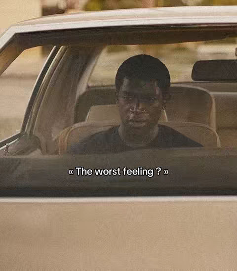 A man is shown sitting in the driver's seat of a car, viewed from outside through the windshield. The interior of the car is visible, with beige seats and the frame of the door. The man has dark skin and short, dark hair, with a serious expression on his face. Text overlayed on the image reads, « The worst feeling? »
