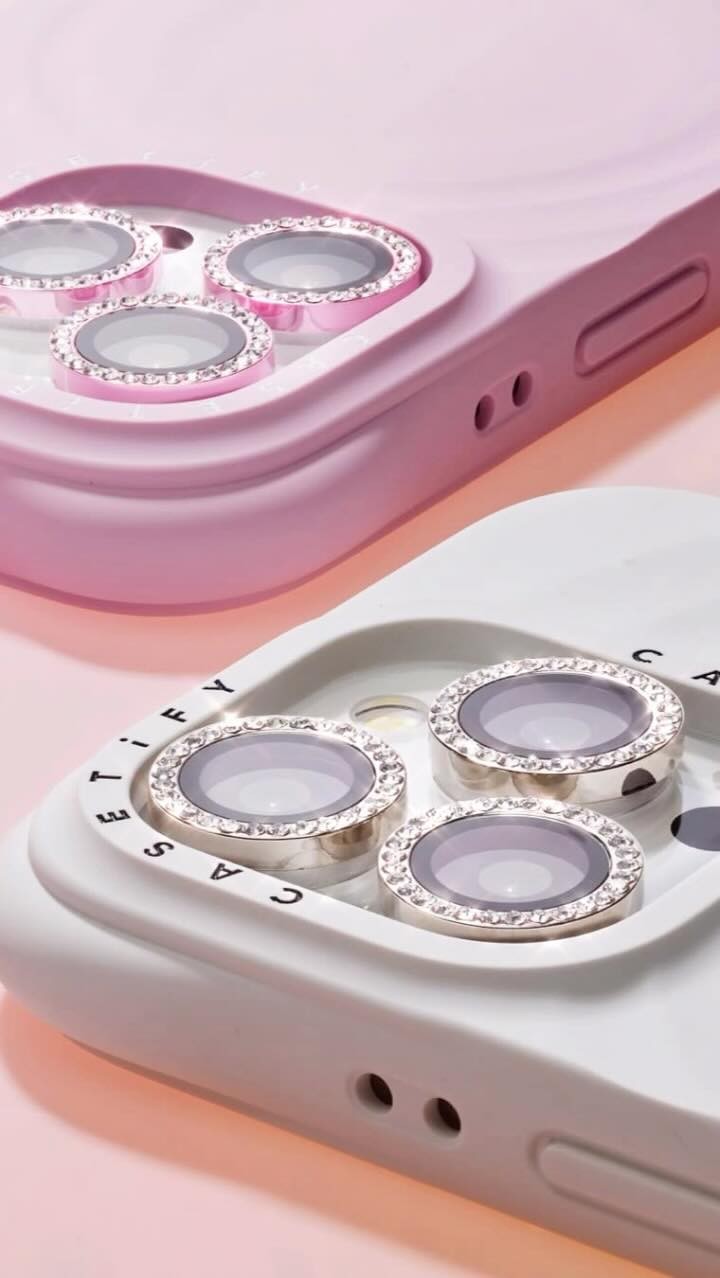 The image shows a close-up of two cell phone cases, one pink and one white. Each case has raised areas designed to protect the camera lenses; the lenses themselves are ringed with silver and encrusted with small, glittering rhinestones or gems. The background fades from light pink to a slightly darker shade.