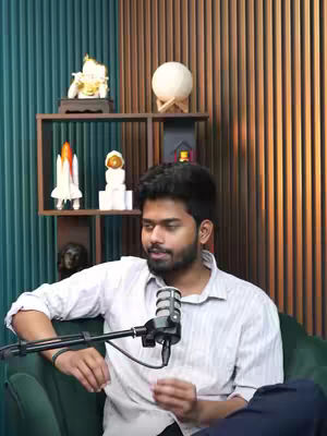 A man in a light-colored shirt sits in front of a wooden and teal wall with shelves holding decorative items, including a golden statue and a model rocket. A microphone is positioned in front of him, and the setup suggests a podcast or video recording environment. The man is looking to his left, and he has a serious expression. The color palette is relatively muted, with shades of teal, brown, and beige.