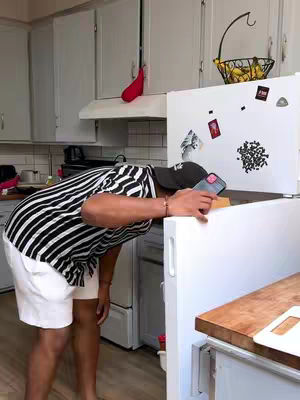 A man stands in a kitchen, leaning over a white refrigerator with a phone in his hand. He wears a striped shirt, white shorts, and a black baseball cap. The kitchen has white cabinets and a wooden countertop. A sign is on the refrigerator. A basket of lemons hangs from a shelf.