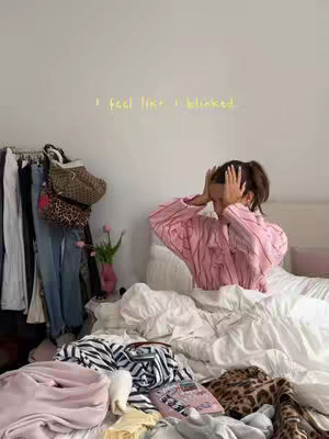 A messy bed with a woman sitting up with her hands on her head. Clothes are scattered on the bed. A rack of clothes can be seen on the left. Text on the wall in yellow cursive says, "I feel like I blinked."
