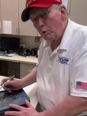 The cover image shows a man, presumably Donald Trump, indoors. He's wearing a white shirt with a "TRUMP" logo and an American flag patch on the sleeve, and a red "Make America Great Again" cap. He is looking at something not visible in the image while holding a pen over a dark object, possibly a book or electronic device, on the table in front of him. The setting appears casual, with office-like elements in the background, including a desk and cabinets.
