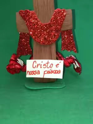 The cover image shows a handmade Easter decoration against a green background. It features a wooden cross adorned with red glitter, a red collar, and two red arm-like structures, with a sign that reads: 'Cristo e nossa páscoa'. The cross sits on a green base.