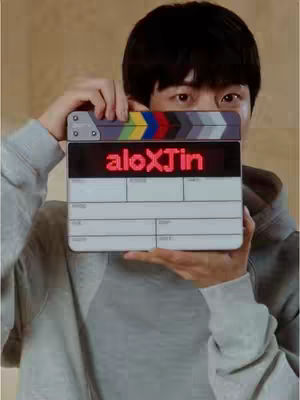 A man with black hair holds a clapperboard in front of his face with the text "aloXJin" displayed. The clapperboard is held so that most of his face, only the eyes and part of forehead are showing. He is wearing a grey hoodie with a neutral background.