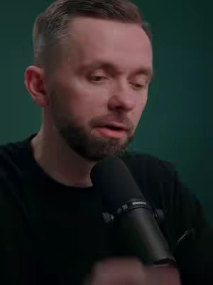 A man with short, neatly styled hair and a trimmed beard is positioned in front of a dark to medium-green background. The man is wearing a black shirt and holding a black microphone, suggesting he is either speaking or about to speak into it. His expression is somewhat serious, and his gaze is directed downward. The lighting appears to be soft, illuminating his face and the microphone well.