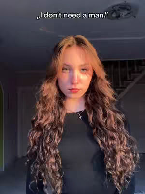 A woman with long, wavy brown hair is centered in the frame. She's wearing a dark-colored top and a silver necklace with a cross pendant. The background is dimly lit with a ceiling fixture visible above her.