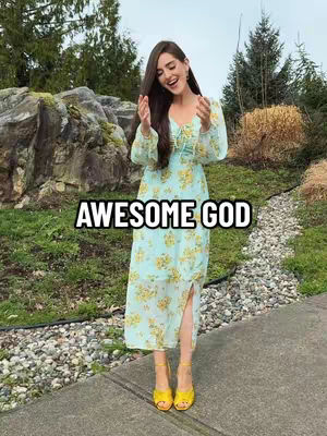 A woman stands outside, hands raised, smiling. She wears a pale blue dress with yellow floral patterns and yellow heels. The text overlayed over her is \"AWESOME GOD\" in white text and a black outline