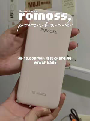 A person's hands hold a white power bank, with text overlaying it. The power bank is rectangular and primarily white. The focus is on the power bank, and the hands holding it. The background includes a light switch, the corner of a wall, and some items attached to the wall. The overall composition is simple, centered around the product.